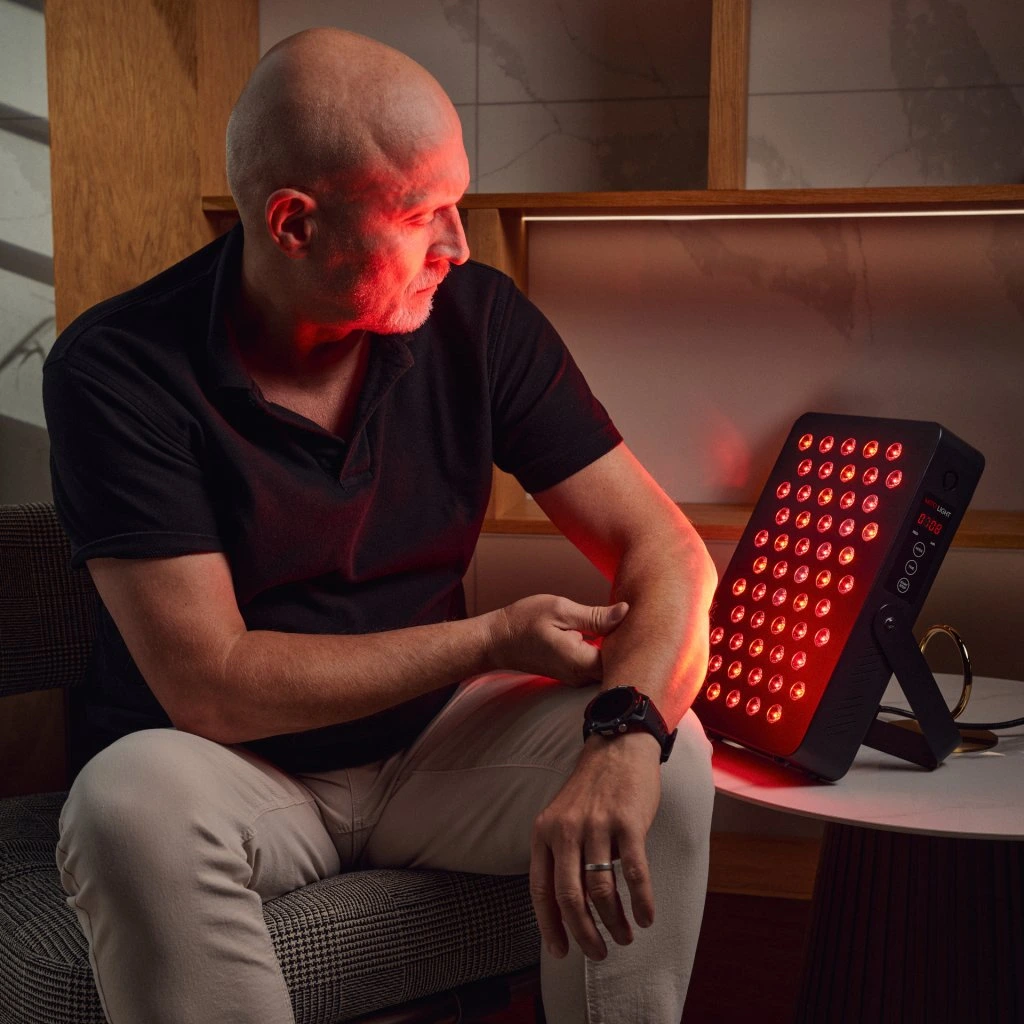 Muž sediaci na kresle ožaruje predlaktie kompaktným červeným LED panelom MitoLight Intro 5.0 na terapiu červeným svetlom