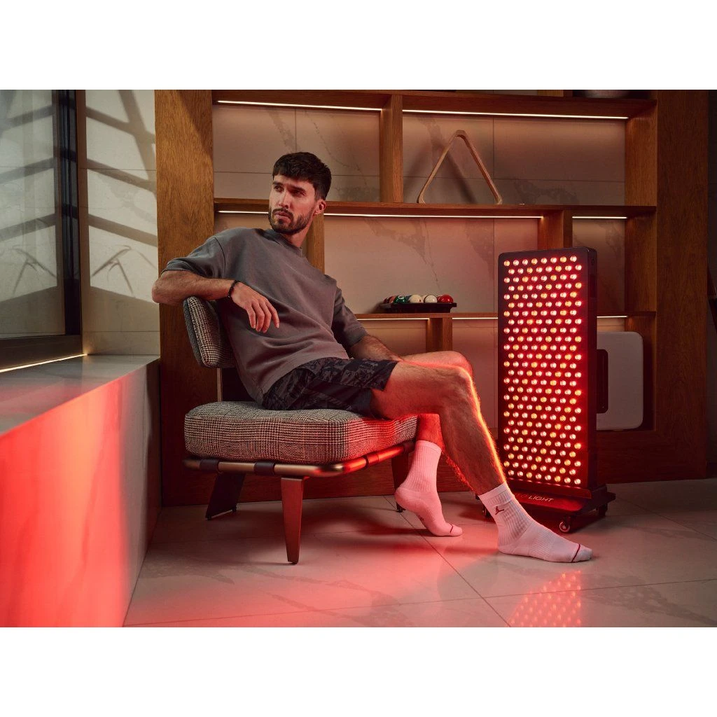stojan-mito-light-floor-stand-5-0-3 Muž sedí na stoličke pri svetelnom paneli MITO LIGHT® na stojane Floor Stand 5.0 – terapia červeným svetlom nôh v sede