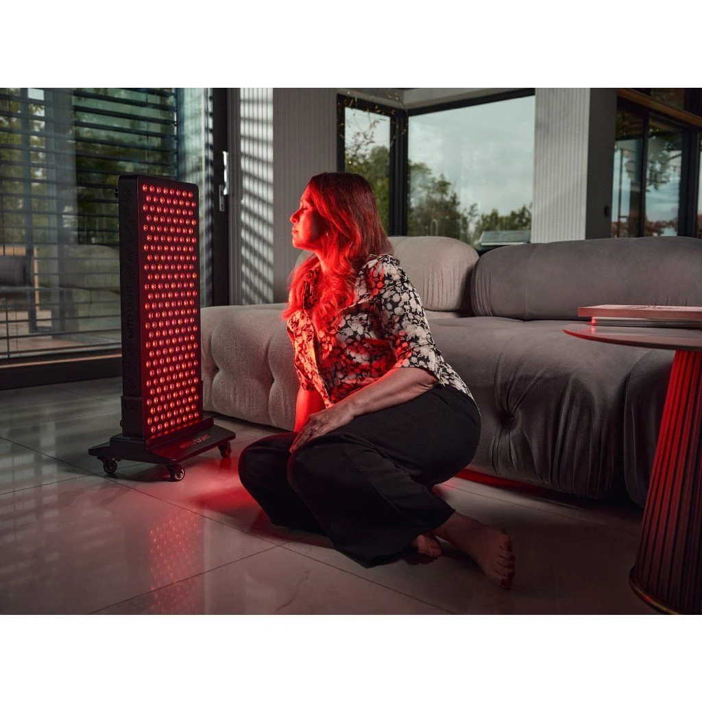 stojan-mito-light-floor-stand-5-0-5 Žena sedí na podlahe pri svetelnom paneli MITO LIGHT® na stojane Floor Stand 5.0 – terapia červeným svetlom chrbta v domácom prostredí